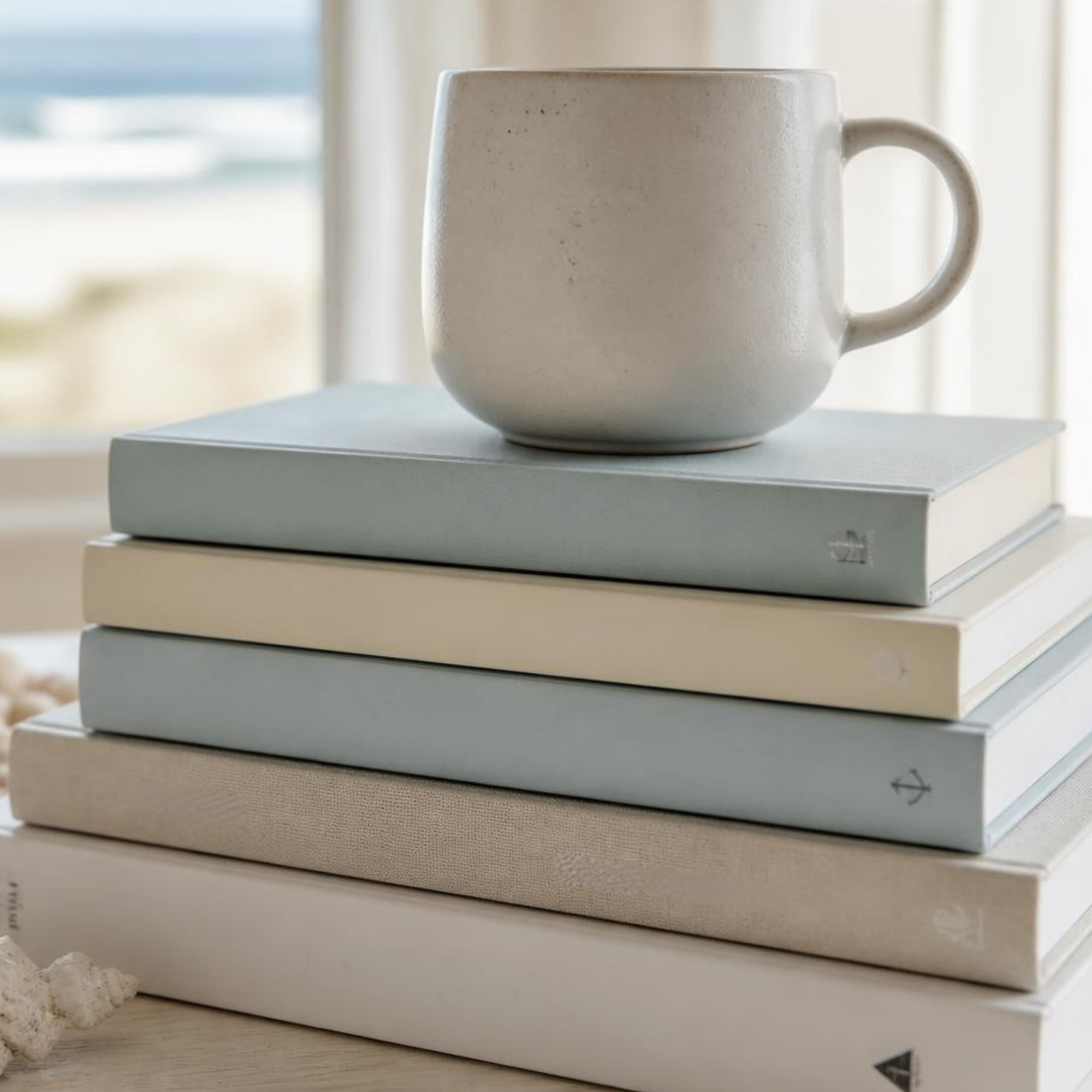 A ceramic mug on a stack of sea-glass blue and cream books by the ocean